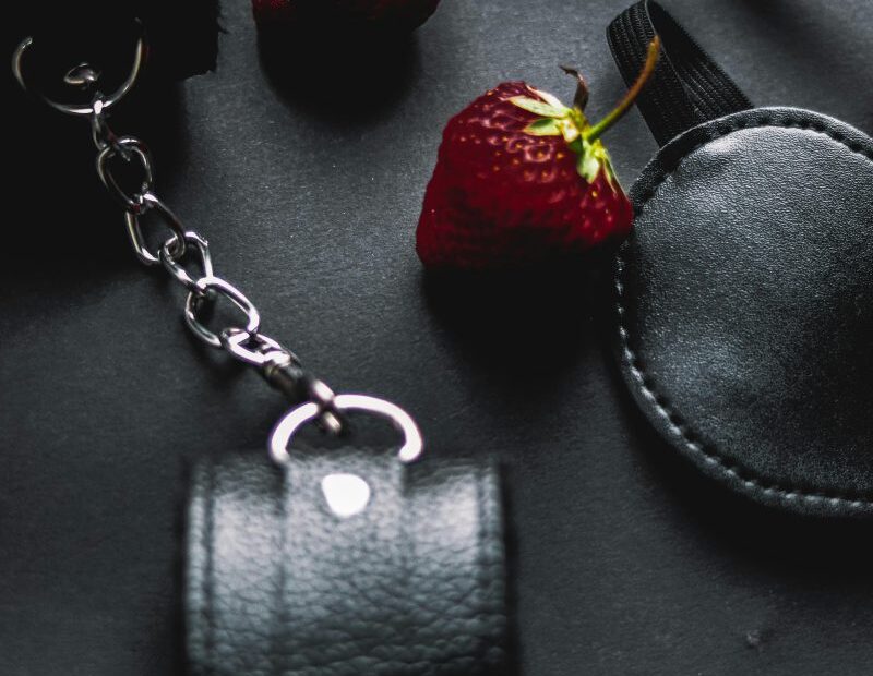 Flat lay of leather BDSM cuffs, paddle and flogger with strawberries on black