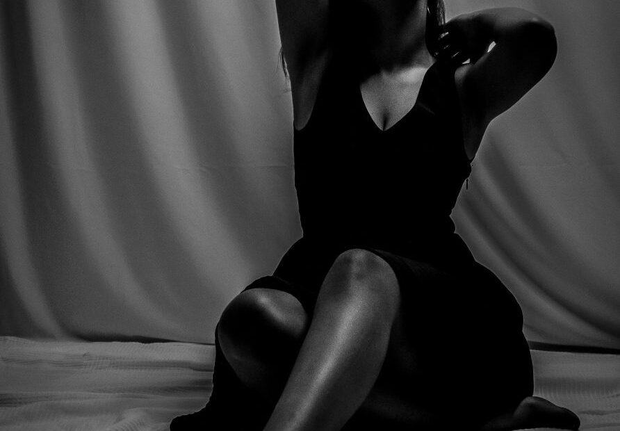 Black and white photo of a woman in a black dress seated on a sheet against draped fabric