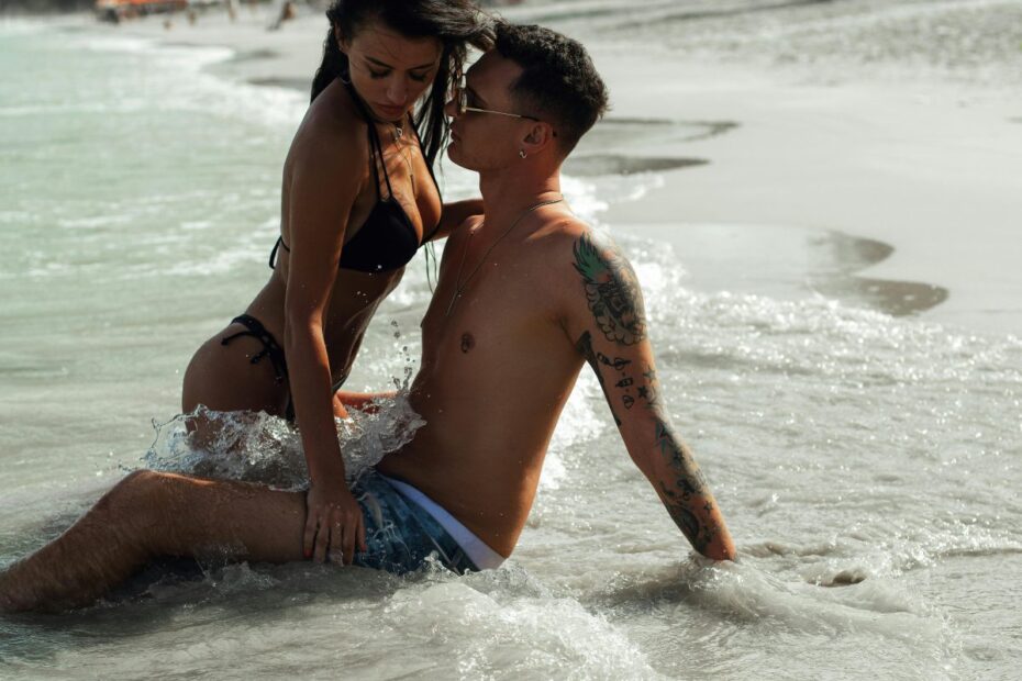 Tattooed couple in swimwear embracing closely while sitting in shallow beach surf