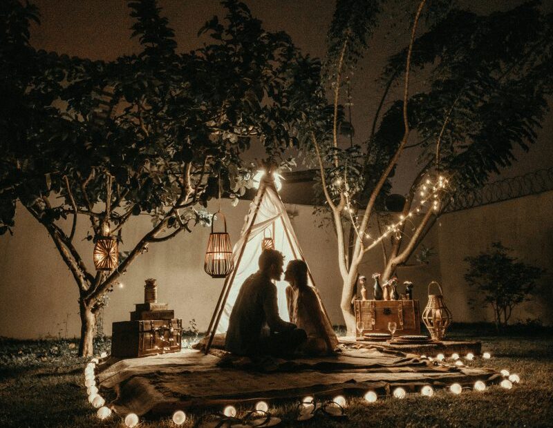 Couple sitting close in a candlelit backyard teepee strung with fairy lights at night