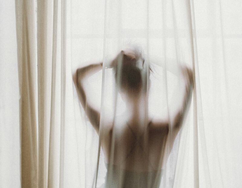 Soft silhouette of a nude woman behind sheer curtains at a sunlit window