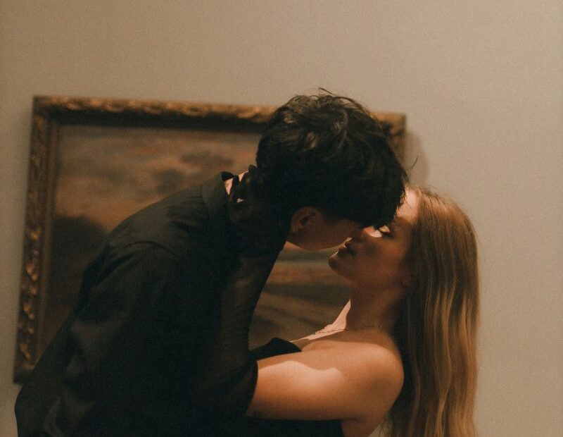 Couple kissing against a wall beneath a framed painting in warm light